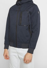 Mörkblå jacka i lättviktsmaterial; har dragkedja fram, elastiska mudd, en stor bröstfick och en texturerad yta.