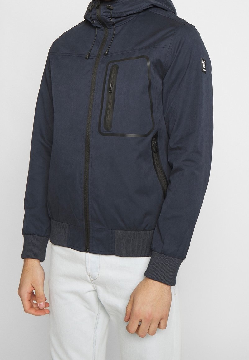 Mörkblå jacka i lättviktsmaterial; har dragkedja fram, elastiska mudd, en stor bröstfick och en texturerad yta.