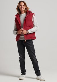 Gilet rembourré rouge avec une capuche et des réglages à cordon, sur un pull texturé gris, associé à un jean noir et des baskets blanches.