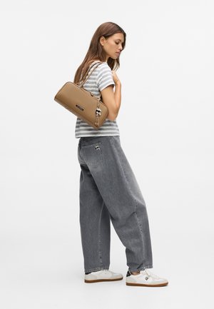 Brun rektangulær håndtaske med en rem, bæres tværs over kroppen. Model i grå lige bukser og en grå-hvid stribet t-shirt, hvide sneakers.