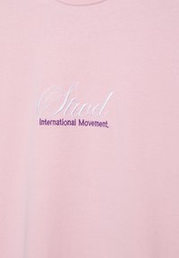 Rosa Stoff mit weiß besticktem kursivem Text "Stud" und lila Text "International Movement" darunter.
