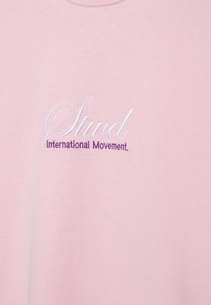 Roza tkanina z vezenim belim kurzivnim napisom "Stud" in vijoličnim napisom "International Movement" spodaj.