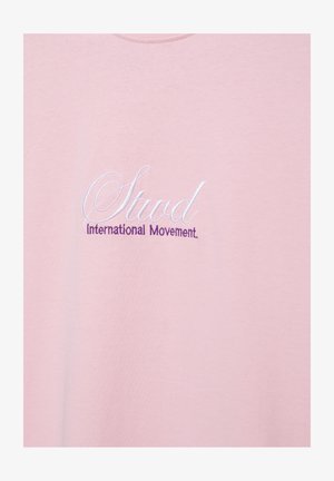 Roza tkanina z vezenim belim kurzivnim napisom "Stud" in vijoličnim napisom "International Movement" spodaj.