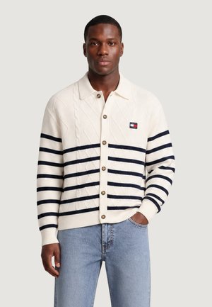 Uomo che indossa un cardigan bianco abbottonato con strisce orizzontali nere e jeans azzurri, in piedi con una mano in tasca.