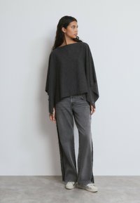 Mujer vistiendo un poncho suelto gris oscuro con mangas anchas, jeans grises de pierna ancha con flecos laterales y zapatillas blancas, de pie frente a una pared lisa.