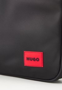 Sort syntetisk taske med en tekstureret overflade og et fremtrædende rødt rektangulært mærke med ordet "HUGO" i sorte bogstaver.