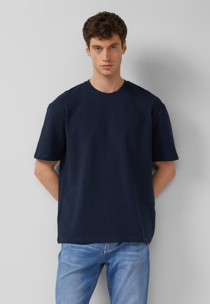 Jonge man met kort krullend haar, gekleed in een marineblauw getextureerd T-shirt met korte mouwen en lichtblauwe spijkerbroek, staand tegen een grijze muur.