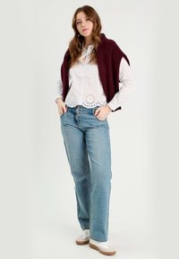 Gilet en tricot bordeaux sur une chemise blanche à boutons avec des détails en œillets, associé à un jean droit bleu clair et des baskets blanches.
