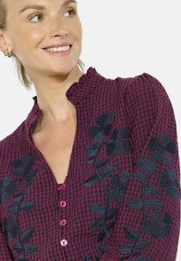 Raudona ir juoda gingham stiliaus bliuska su gėlių siuvinėjimu, raukšlėta apykakle ir rožinėmis mygtukėmis. Audinys turi minkštą tekstūrą ir prigludusią siluetą.