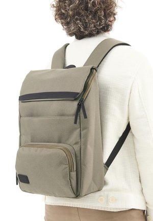 Person, der eine hellbeige Jacke und braune Hosen trägt, mit einem großen olivgrünen Rucksack, der mehrere Reißverschlussfächer hat.