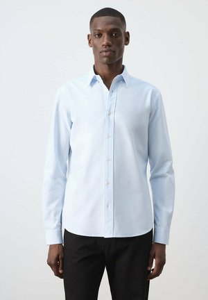 Hombre con camisa de manga larga azul claro abotonada y pantalones negros, de pie contra un fondo blanco sencillo.