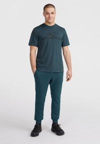 Turkusowa koszulka z czarnym logo, połączona z dopasowanymi turkusowymi dresami i czarnymi butami. Posiada krótkie rękawy i luźny krój.