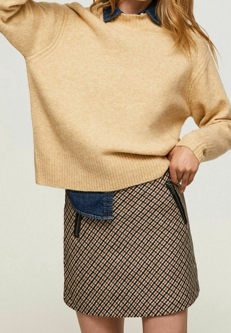 Maglione beige con orlo a costine, abbinato a una gonna a fantasia con linee diagonali marroni, nere e crema e dettagli in denim sulle tasche.