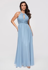 Femme portant une robe longue bleu clair à col halter avec un corsage plissé et un décolleté en forme de trou de serrure, posant avec une main sur la hanche.