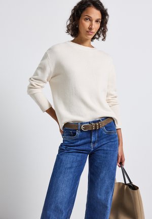 Pull en maille crème avec un col rond, associé à un jean bleu à jambes larges et une ceinture brune. La personne tient un sac fourre-tout beige.