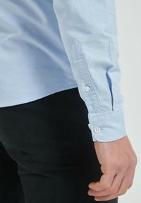 Chemise à manches longues bleu clair avec un poignet boutonné. Design ajusté et texture lisse, présentée avec un pantalon noir en arrière-plan.