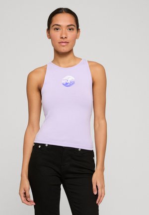 Žena oblečená v bezrukávovom levanduľovom topu s bielym a modrým vlnovým logom a čiernymi džínami, stojaca pred jednoduchým svetlým pozadím.