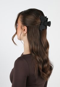 Fermaglio per capelli a forma di fiocco nero fissato in capelli scuri e mossi, con una finitura liscia e una superficie texturizzata. Il fermaglio è posizionato vicino alla parte posteriore della testa.
