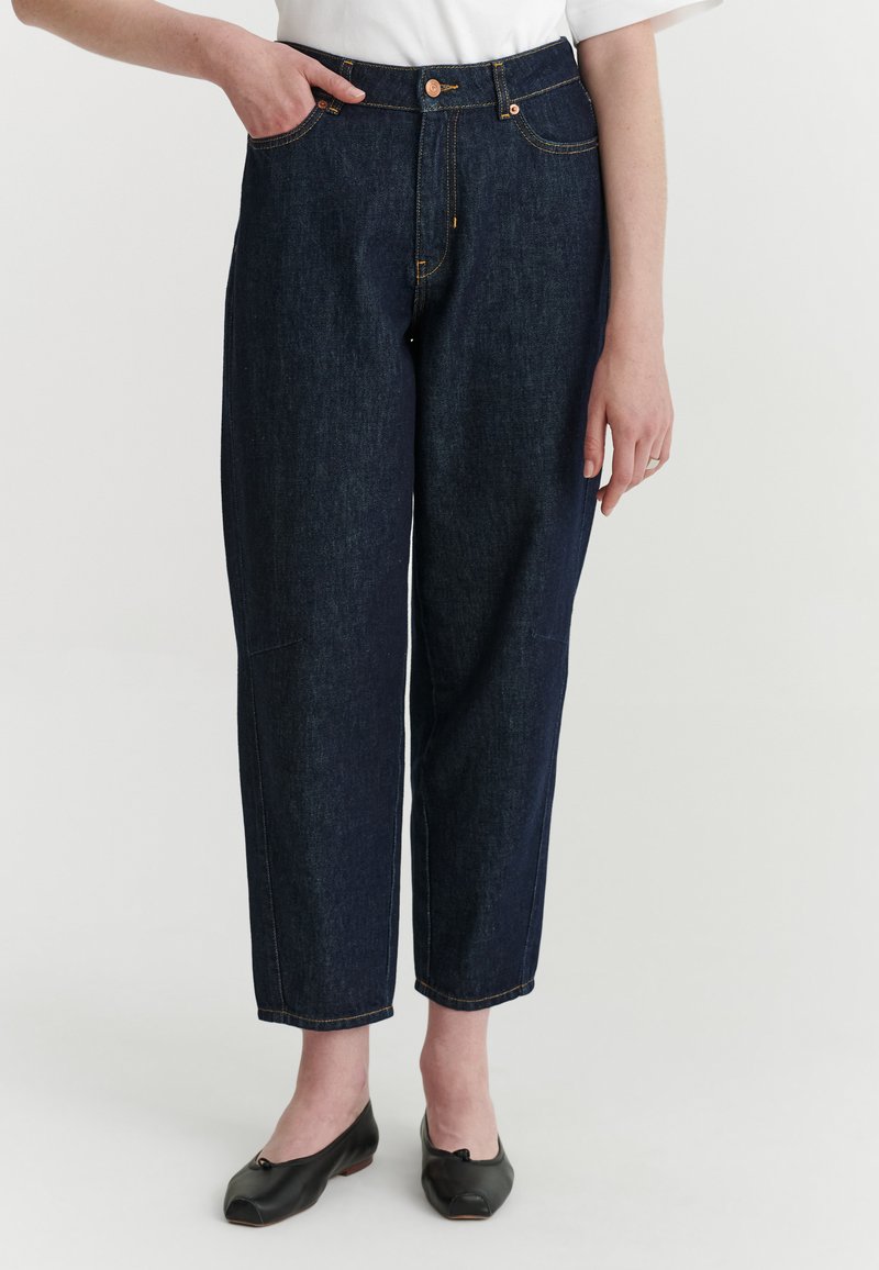 Personne portant un jean droit bleu foncé, un haut blanc et des chaussures plates noires, debout avec une main dans la poche, sur un fond uni.