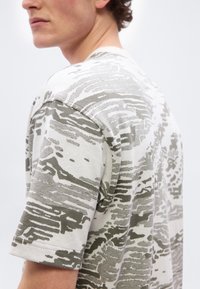 Νεαρός άνδρας που φοράει ανοιχτόχρωμο πουκάμισο μ ε κοντά μανίκια και γκρι αφηρημένο καμουφλάζ σχέδιο, απεικονίζεται από τον ώμο έως το μέσο του στήθους, στραμμένος πλάγια.