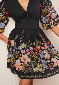Robe noire avec broderies florales présentant des fleurs colorées. Manches courtes bouffantes et un lien au col. Jupe évasée.