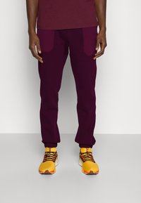 Bordeauxfarbene Cargohosen mit zwei Fronttaschen, schmal zulaufenden Bündchen und einem elastischen Bund, kombiniert mit gelben Sneakers, die schwarze Schnürsenkel und orangefarbene Akzente haben.