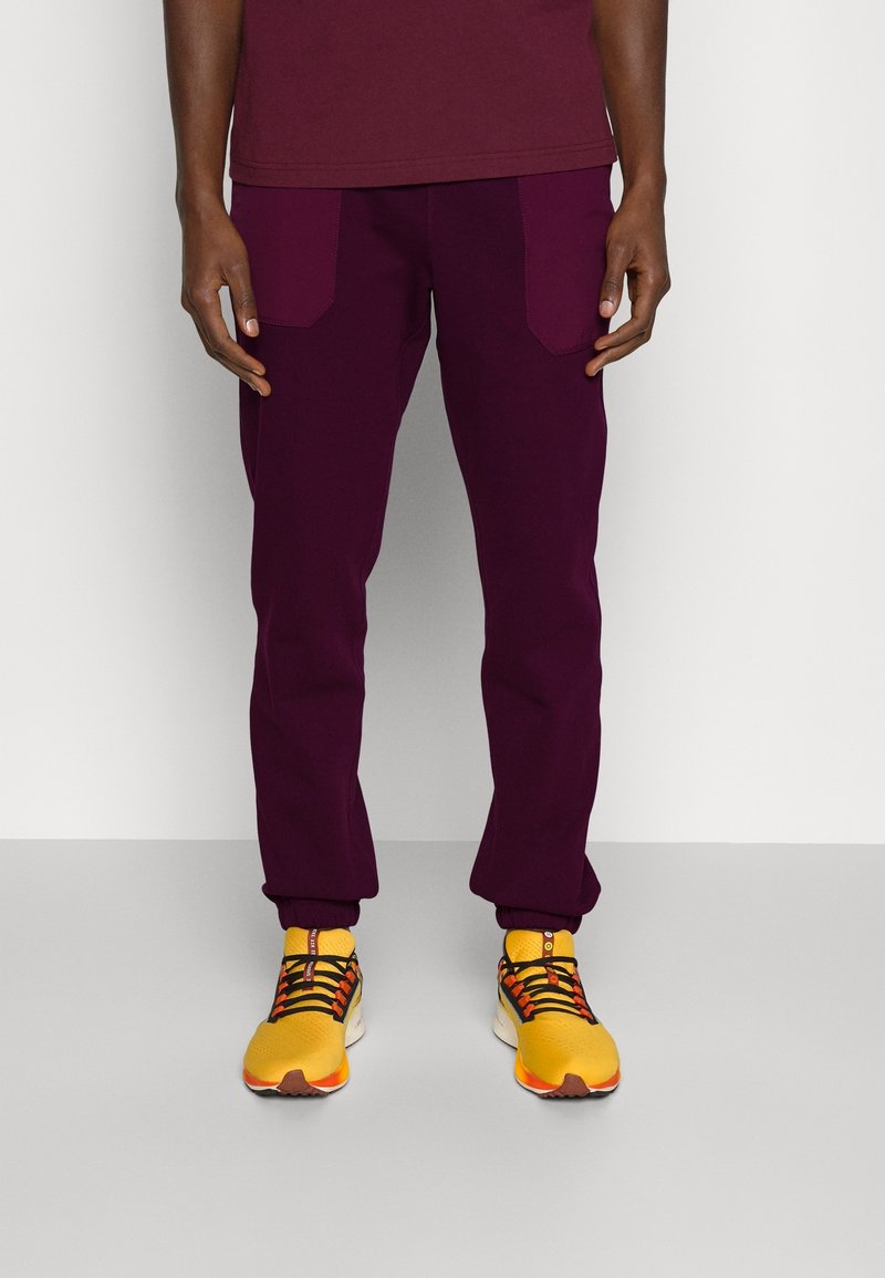 Bordeauxfarbene Cargohosen mit zwei Fronttaschen, schmal zulaufenden Bündchen und einem elastischen Bund, kombiniert mit gelben Sneakers, die schwarze Schnürsenkel und orangefarbene Akzente haben.