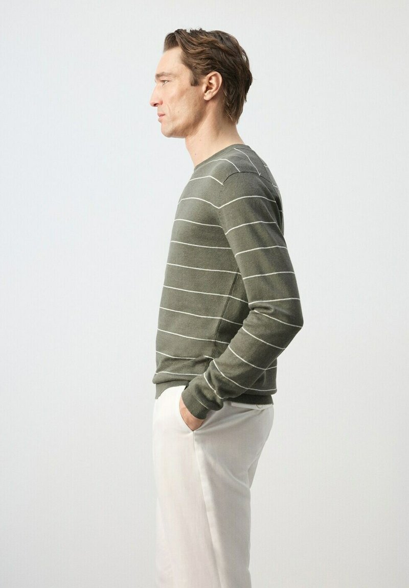 Homme en chemise à manches longues rayée vert et pantalon blanc, debout de profil, les mains dans les poches, sur un fond clair uni.