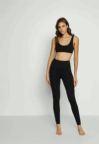 Jovem mulher em pé descalça contra uma parede simples, vestindo um top cropped preto e leggings pretas de cintura alta.