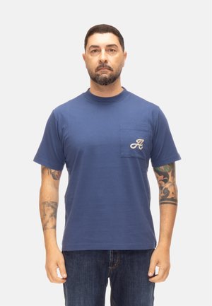Uomo con capelli corti e scuri e barba, indossa una maglietta blu con taschino e la lettera "A", in piedi davanti a uno sfondo chiaro e uniforme. Tatuaggi sugli avambracci.