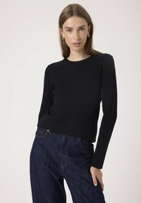 Black ribbed knit sweater with long sleeves and a round neckline, paired with dark denim jeans. Sweater has a fitted silhouette and textured fabric.