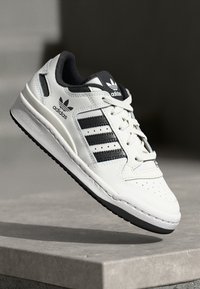 Chaussure de sport blanche avec des rayures et des accents noirs, fabriquée en cuir et en maille. Présente une pointe perforée et une semelle extérieure en caoutchouc pour une meilleure adhérence.