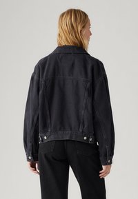 Črna jeans jakna z ohlapnim krojem, velikim ovratnikom in z gumbi zaprtimi manšetami. Ima srebrne dodatke in ravno šivanje na hrbtni strani.