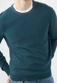Mann trägt einen dunkelgrünen Pullover mit Rundhalsausschnitt über einem weißen T-Shirt, kombiniert mit blauen Jeans, vor einem schlichten weißen Hintergrund.