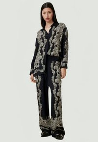 Γυναίκα που φοράει μαύρο σετ με ασύμμετρη μπλούζα και παντελόνι φαρδύ, με περίπλοκα μπεζ μοτίβα paisley, στέκεται μπροστά σε ουδέτερο φόντο.