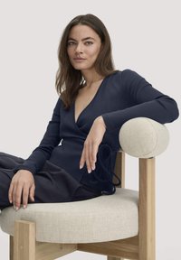 Vrouw met lang bruin haar, gekleed in een marineblauwe omslagtrui en donkere broek, ontspannen zittend op een beige, met kussens beklede houten stoel.