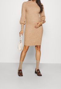 Robe longue en maille à manches longues, beige, avec texture côtelée et silhouette ajustée. Associée à des chaussettes beiges et des chaussures marron ; tenant un sac à main blanc.