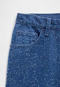 Denimfarkut keskitummassa sinisessä sävyssä, jossa on pistekuvioinen tekstuuri, etutasku ja metallinen nappikiinnitys.