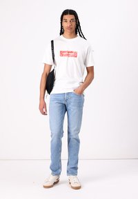 Hvid t-shirt med rødt logo, lyseblå jeans og hvide sneakers. En sort skuldertaske hænger over venstre skulder.