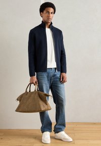 Ναυτική ζακέτα με φερμουάρ, λευκό πουκάμισο, μπλε τζιν και λευκά αθλητικά παπούτσια. Καφέ δερμάτινη τσάντα με δύο χερούλια, στρογγυλή μορφή και μαλακή υφή.