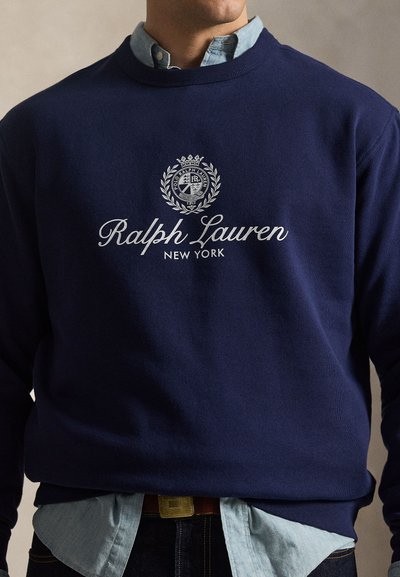 Άνδρας που φοράει μπλε σκούρο φούτερ Ralph Lauren πάνω από ανοιχτό μπλε πουκάμισο με κουμπιά, με τζιν και καφέ ζώνη.