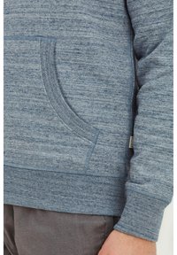 Sweatshirt bleu mélangé avec une poche avant, des poignets et un ourlet côtelés, et un tissu texturé, montré partiellement sur un bras.