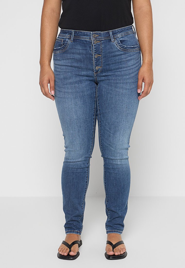 Schlanke Jeans aus mittelblauem Denim mit einem Fünf-Knopf-Verschluss, vorderen Taschen und gesteppten Akzenten. Getragen mit schwarzen Flip-Flops.