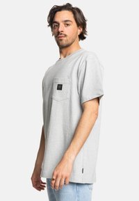 Grå bomulds t-shirt med korte ærmer, en rund halsudskæring og en venstre brystlomme med et sort logo-patch. Side slids-detaljer ved kanten.