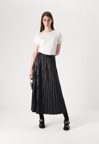 Jupe maxi plissée noire avec une taille noire, associée à un t-shirt blanc. Le mannequin porte un petit sac à main blanc et des chaussures noires.