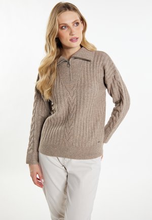 Donna con lunghi capelli biondi che indossa un maglione lavorato a trecce color taupe con collo a mezza zip e pantaloni chiari, in piedi davanti a uno sfondo semplice.