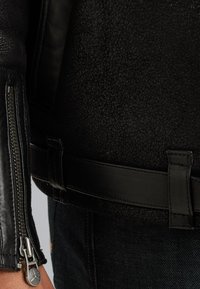 Zwarte leren jas met een gestructureerde afwerking, ritsmanchetten en een design met ceintuur in de taille, aangevuld met soepele accenten.