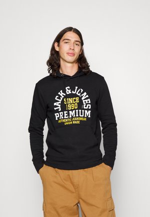 Sudadera negra con texto blanco y amarillo, capucha con cordones y bolsillo frontal. El modelo lleva pantalones cargo beige. Diseño sencillo y casual.