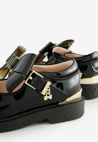 Scarpe in pelle verniciata nera con suola spessa, caratterizzate da un grande fiocco, dettagli dorati e una fibbia, esaltate da un piccolo charm.