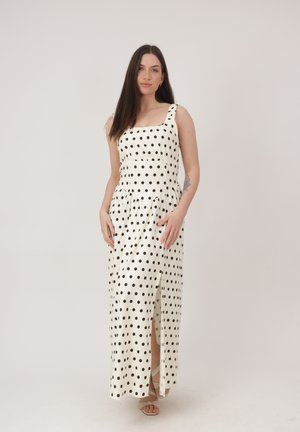 Donna con capelli lunghi e scuri che indossa un maxi abito bianco senza maniche con pois neri e spacco frontale, in piedi contro uno sfondo neutro.